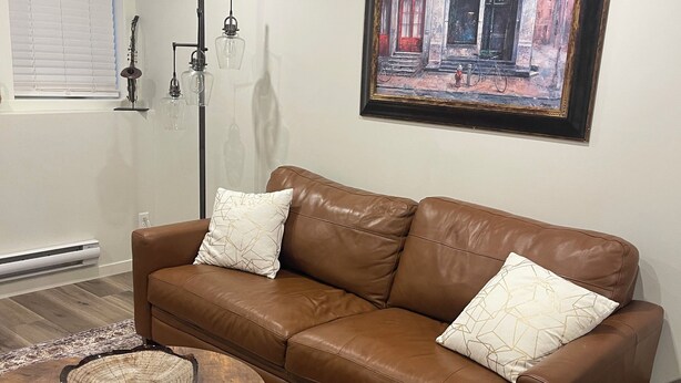 A living room with a brown leather sofa, two patterned cushions, a round wooden coffee table, a patterned rug, a painting on the wall, and a floor lamp. - Adorable 1-bedroom condo in beautiful Calgary perfect for your stay (Calgary)