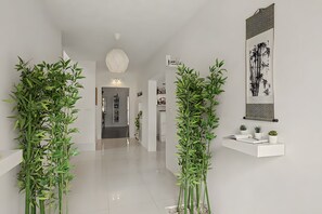A hallway with bamboo plants, a white shelf with books and small plants, and a wall with a framed artwork.