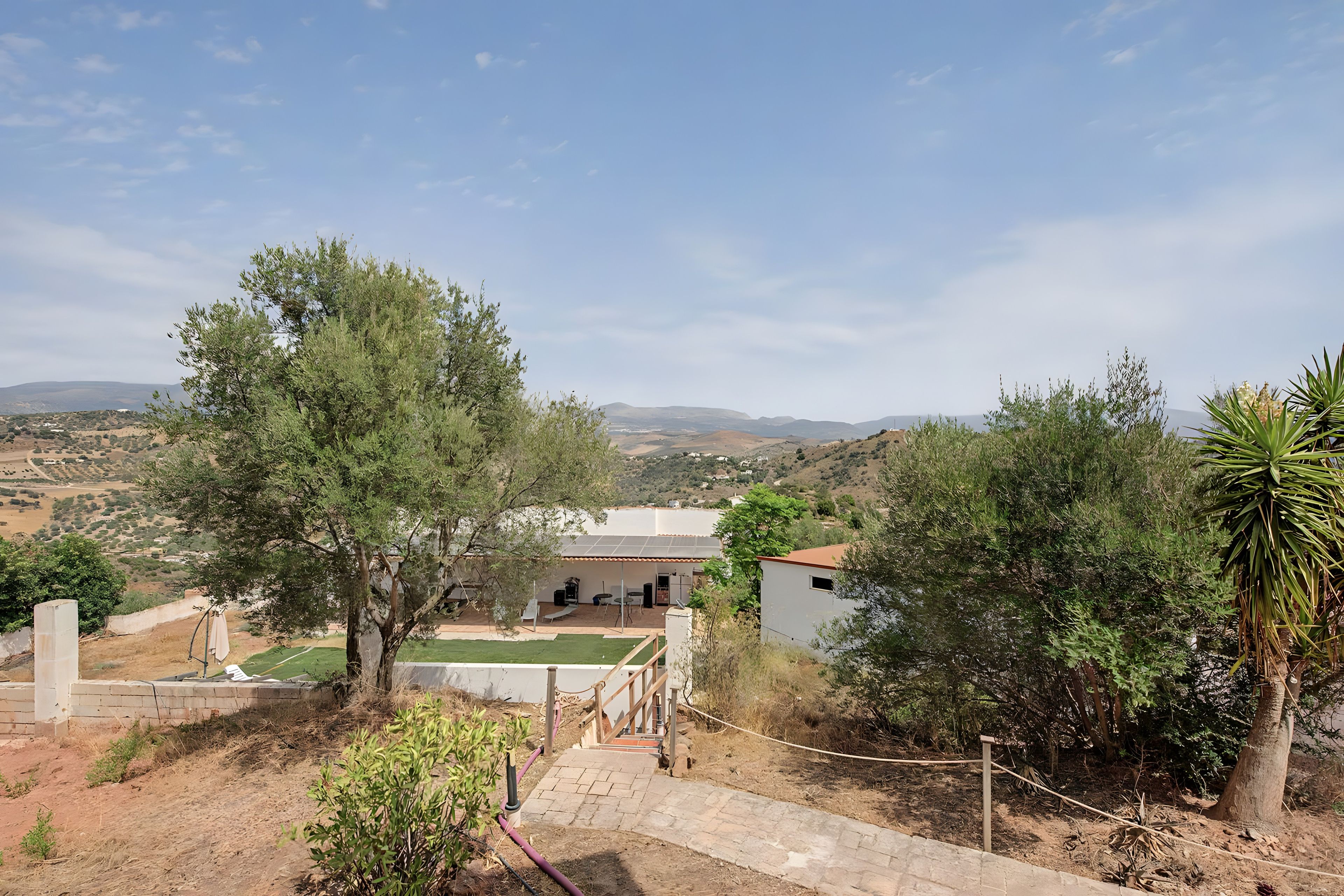 Una propiedad residencial con un camino pavimentado que lleva a una casa con paneles solares en el techo, rodeada de árboles y un paisaje montañoso.