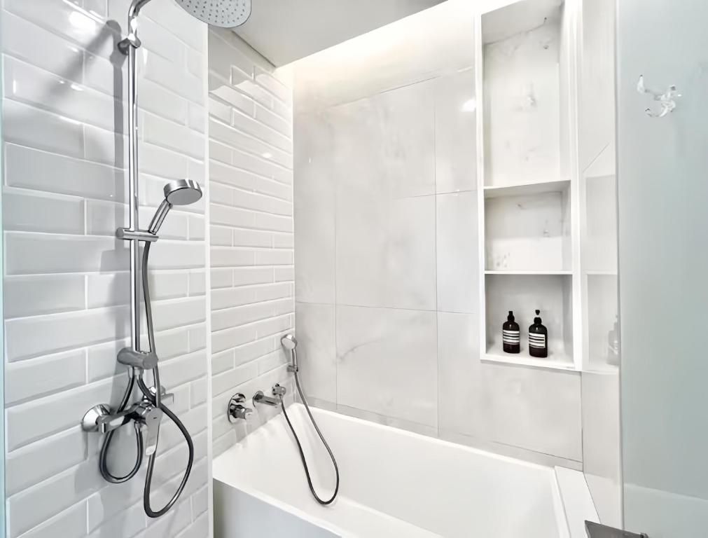 A modern bathroom with a white bathtub, a handheld showerhead, and a wall-mounted faucet. There are white tiled walls and a white shelf with toiletries.