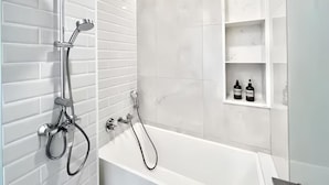 Et moderne badeværelse med et hvidt badekar, et håndholdt brusehoved og en vandhane fastgjort på væggen. Væggene er beklædt med hvide fliser, og der er en hvid hylde med toiletartikler.