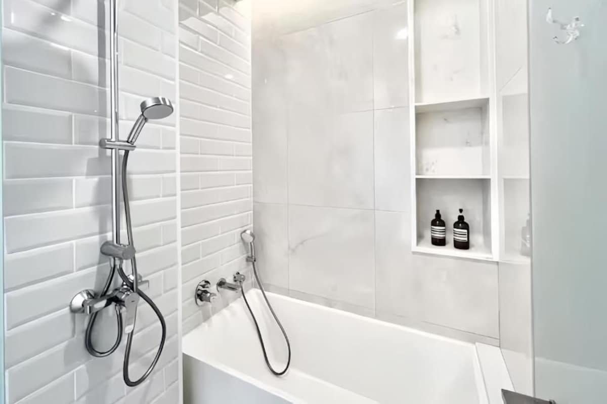 A modern bathroom with a white bathtub, a handheld showerhead, and a wall-mounted faucet. There are white tiled walls and a white shelf with toiletries.