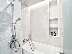 A modern bathroom with a white bathtub, a handheld showerhead, and a wall-mounted faucet. There are white tiled walls and a white shelf with toiletries. - Siamese Residence Sukhumvit 42 Bangkok (Krung Thep Maha Nakhon)