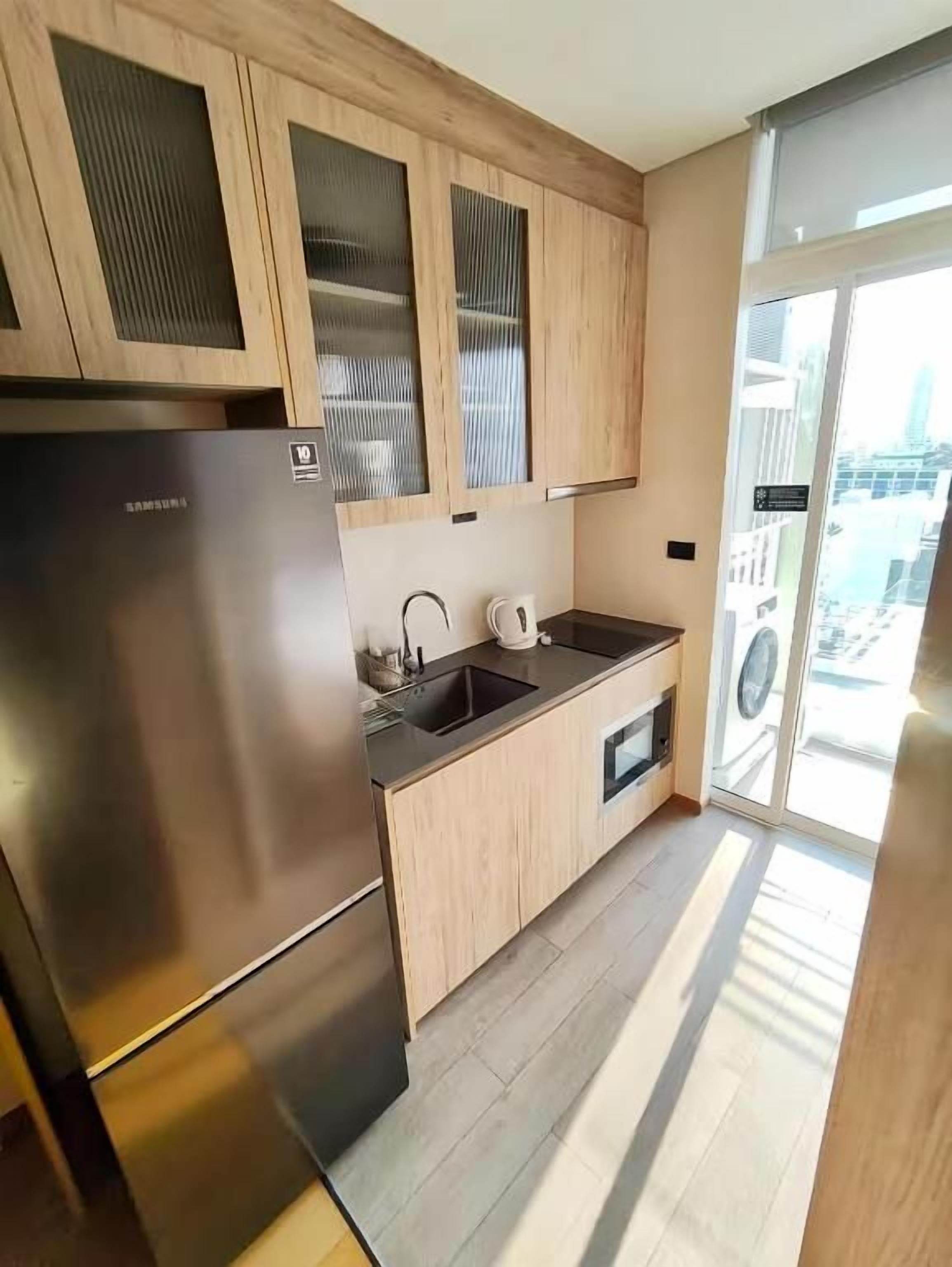 A modern kitchen with a stainless steel refrigerator, wooden cabinets, a sink, and a window providing a view of the city.