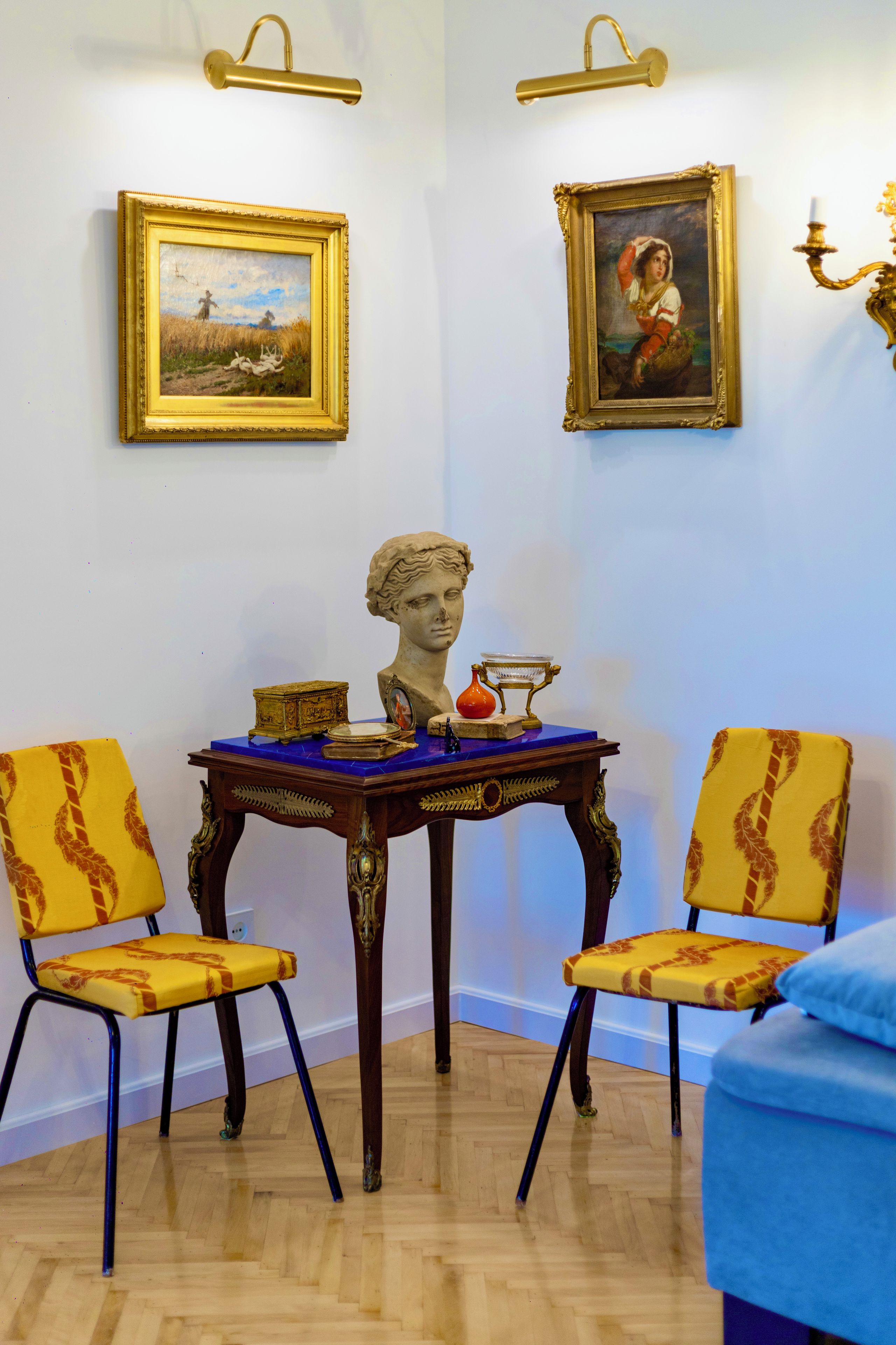 A room with two chairs, a table with a bust, and two framed paintings on the wall.