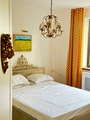 A bedroom with a bed, a chandelier, a painting, and a window with curtains.