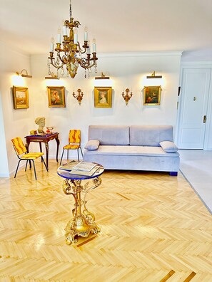 A room with a wooden parquet floor, a blue sofa, a dining table with chairs, a chandelier, and framed artwork on the walls.