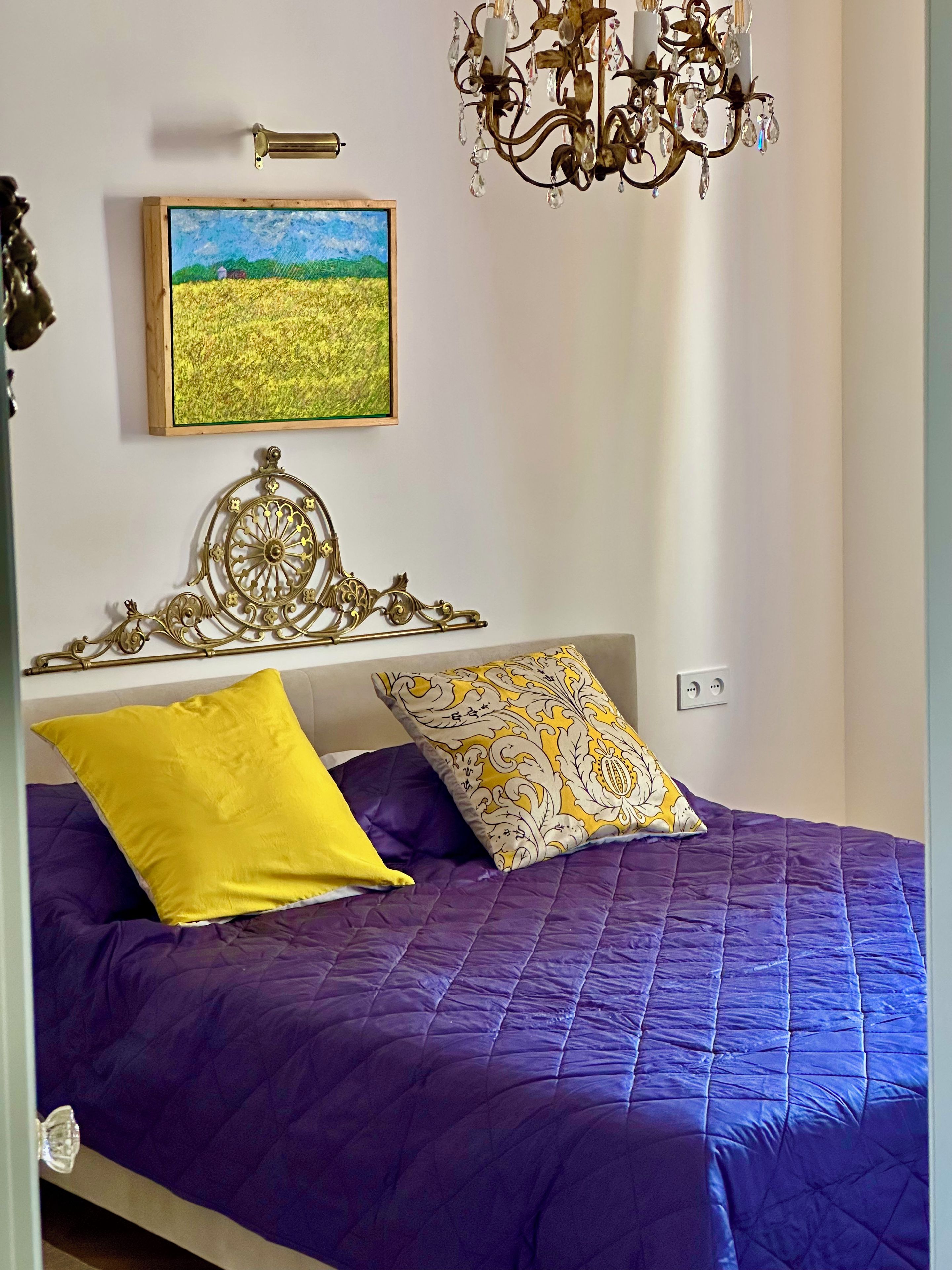 Un dormitorio con colcha morada, almohada amarilla y almohada estampada. Un cuadro de un campo está en la pared, y un candelabro cuelga sobre la cama.