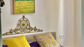 Un dormitorio con colcha morada, almohada amarilla y almohada estampada. Un cuadro de un campo está en la pared, y un candelabro cuelga sobre la cama.