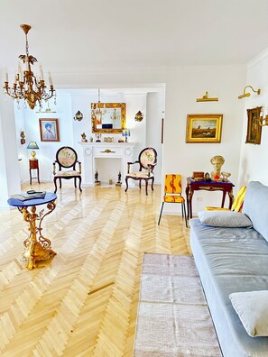 Huoneessa on kalanruotokuviollinen puulattia, sininen sohva, kultainen, koristeellinen pöytä ja kattokruunu.