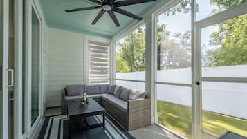 A covered patio area with a ceiling fan, a sectional sofa, a coffee table, and a view of a white fence and greenery.