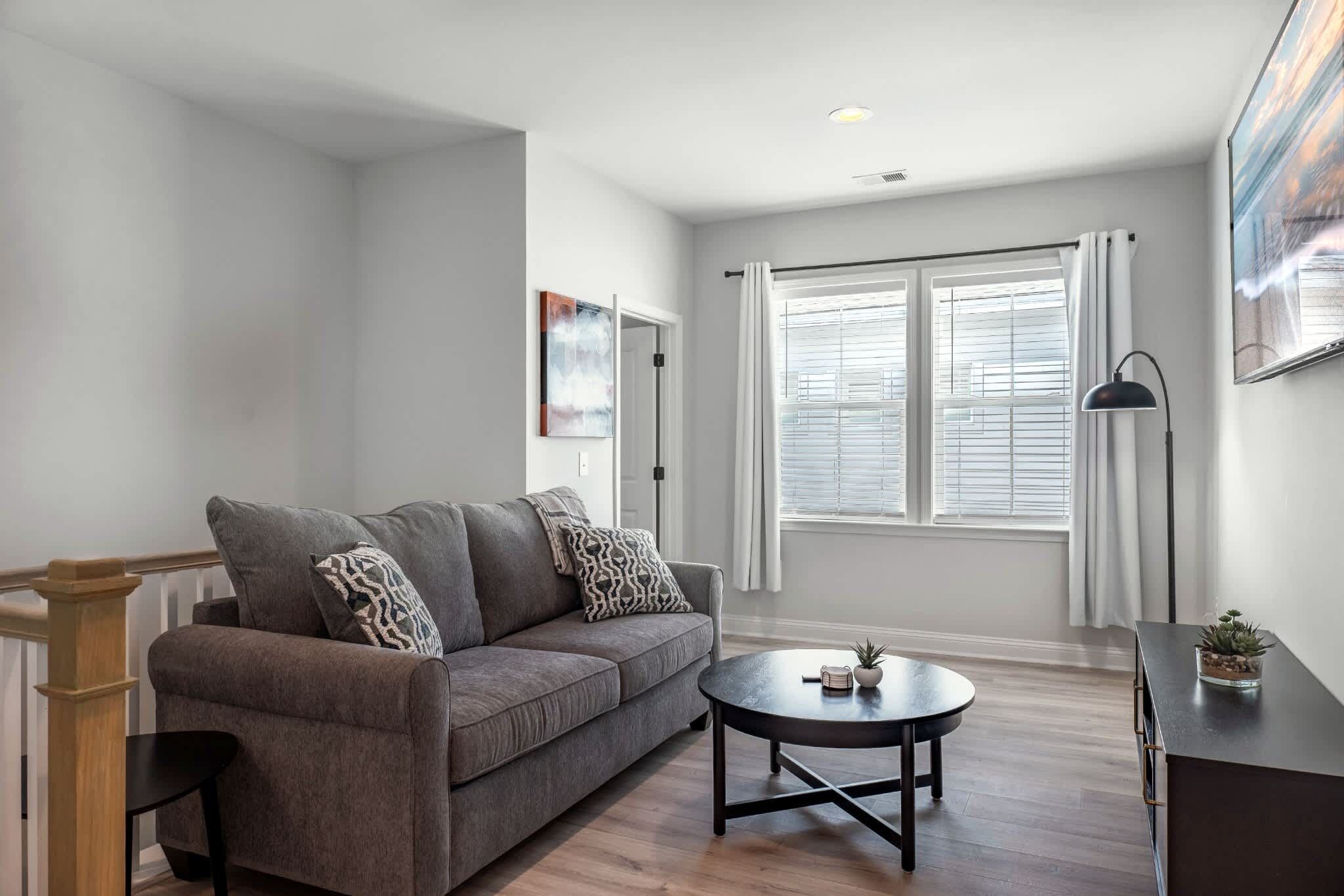 A modern living room with a grey sofa, a round coffee table, and a large window with blinds.