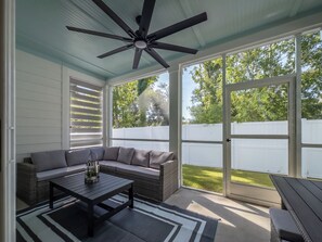 A covered patio with a ceiling fan, a gray sofa, a coffee table, and a view of a white fence and trees.