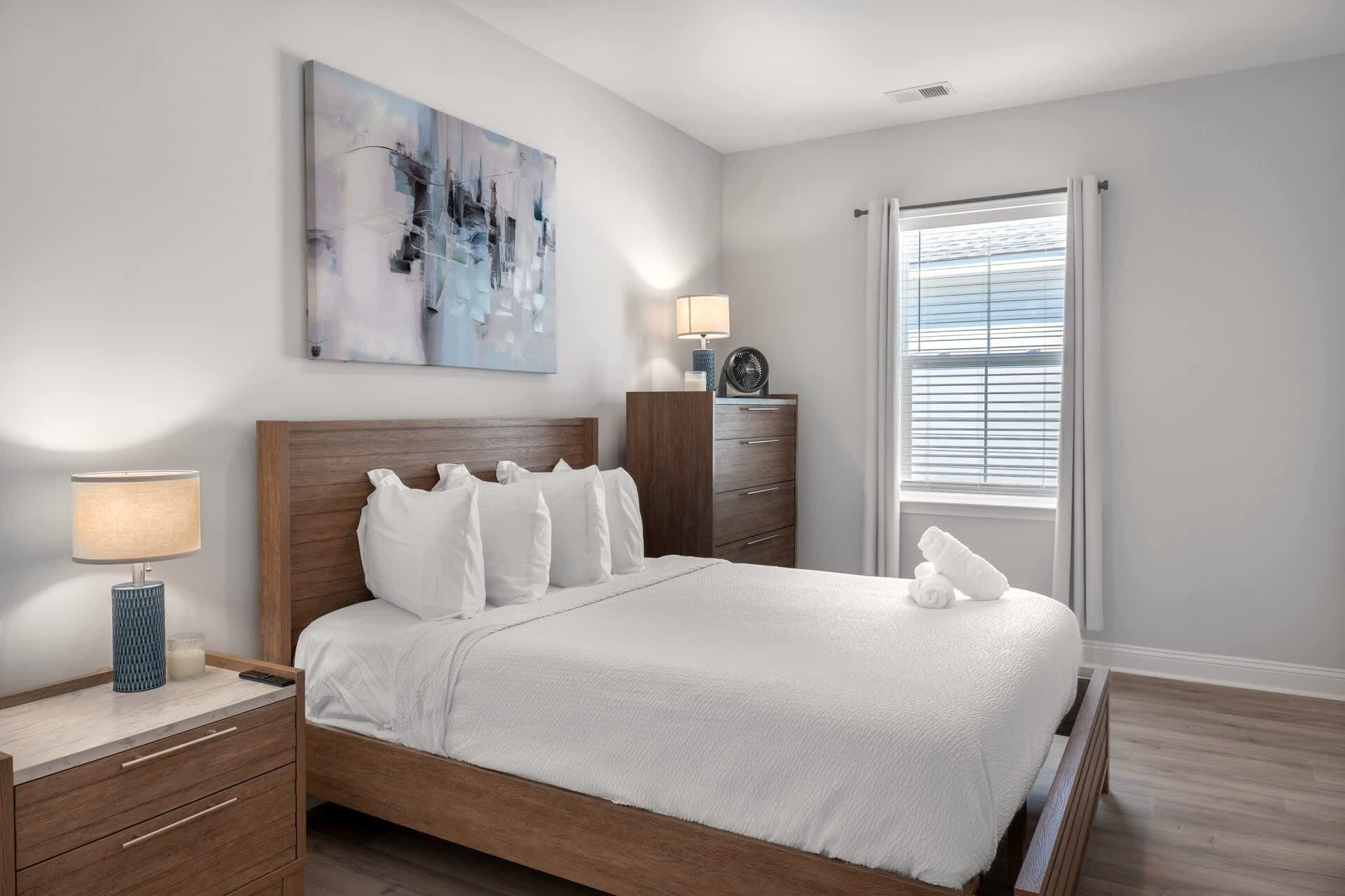 A modern bedroom with a wooden bed, white bedding, a bedside table with a lamp, a dresser, and a large abstract painting on the wall.