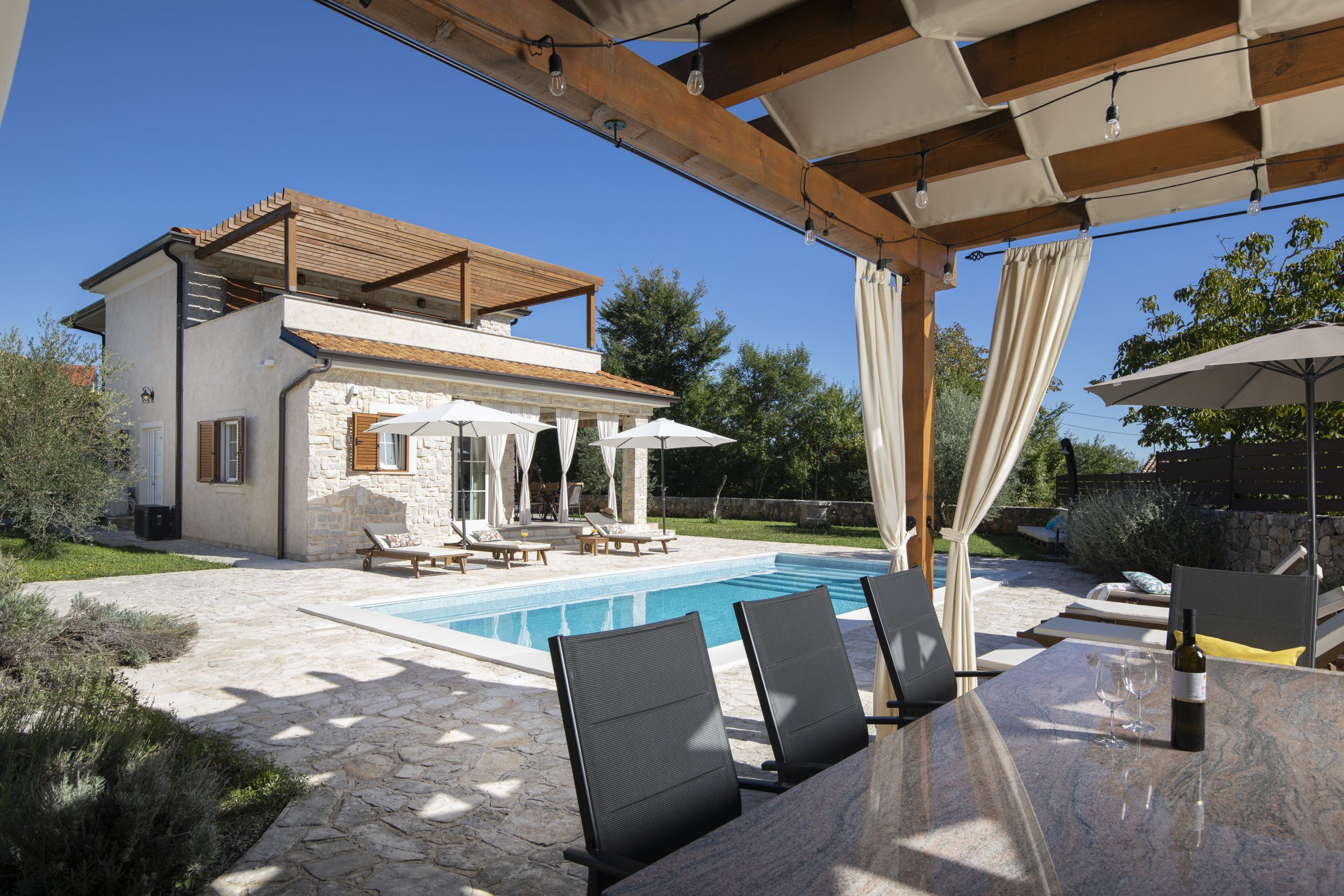 A poolside area with lounge chairs, a table, and a pergola with string lights.