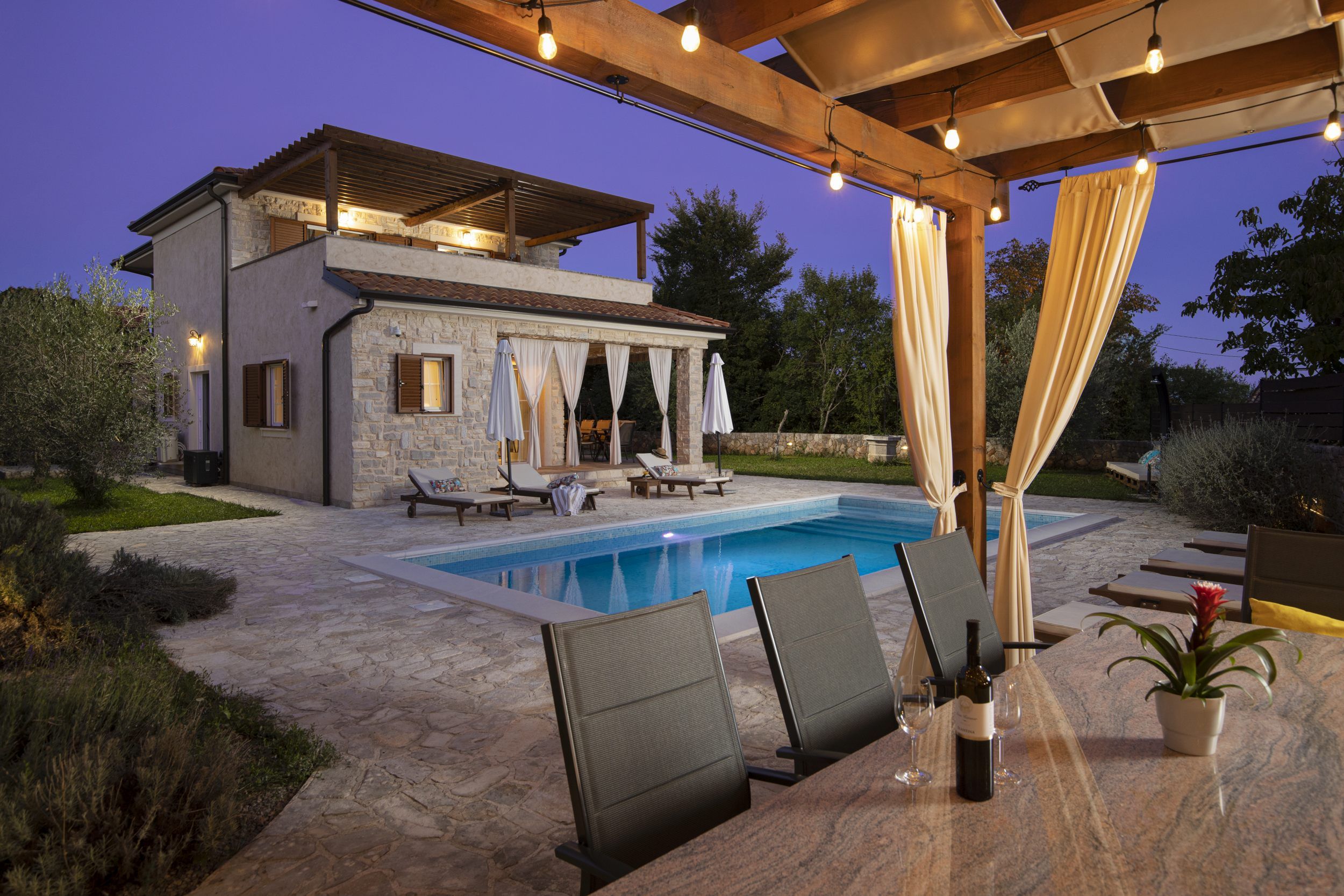 Ein Poolbereich mit Liegestühlen, einer Pergola mit Lichterketten und einem Gebäude aus Stein mit Fenstern.