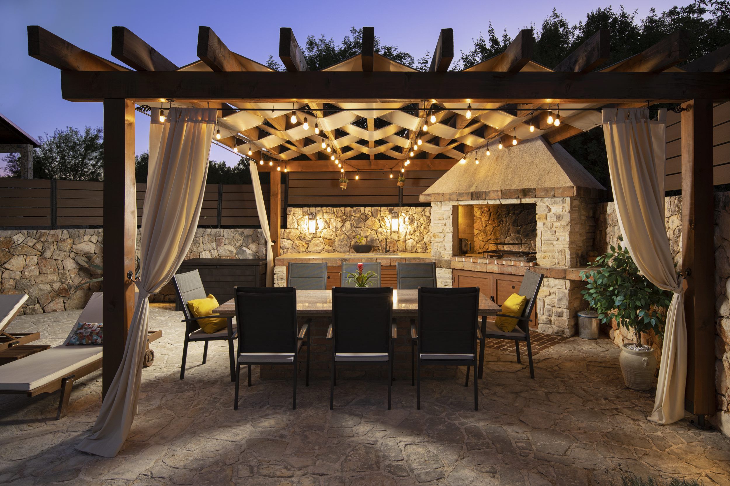 An outdoor patio with a stone fireplace, wooden pergola, and string lights.