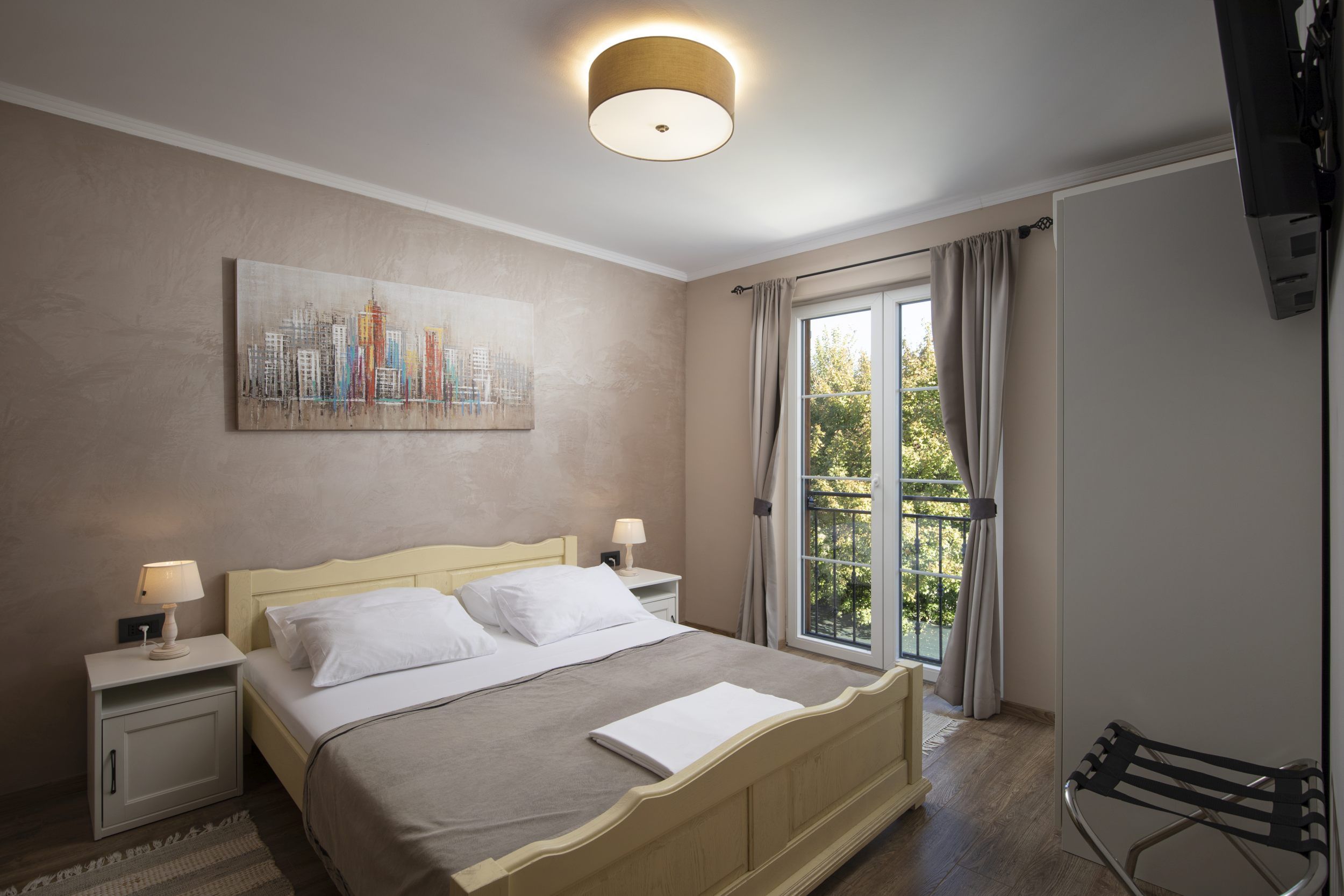 A hotel room with a bed, two bedside tables with lamps, a painting on the wall, a window with curtains, and a chair.