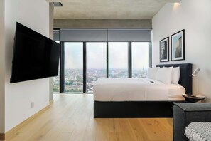 Una habitación de hotel moderna con una cama grande, un televisor de pantalla plana montado en la pared y vistas a la ciudad a través de ventanales del suelo al techo.