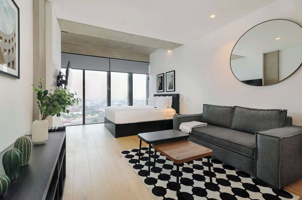 Un dormitorio moderno con una cama grande, un sofá gris, un espejo redondo y vistas a la ciudad a través de amplias ventanas.