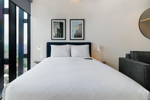 Habitación de hotel con una cama grande, dos cuadros en la pared, un espejo redondo y vistas a la ciudad a través de la ventana.