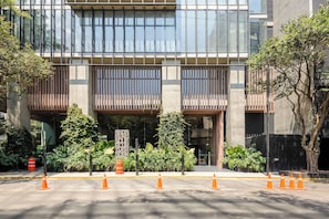 Un edificio moderno con grandes ventanales, un acceso cubierto y un área ajardinada con plantas y conos de tráfico naranjas.