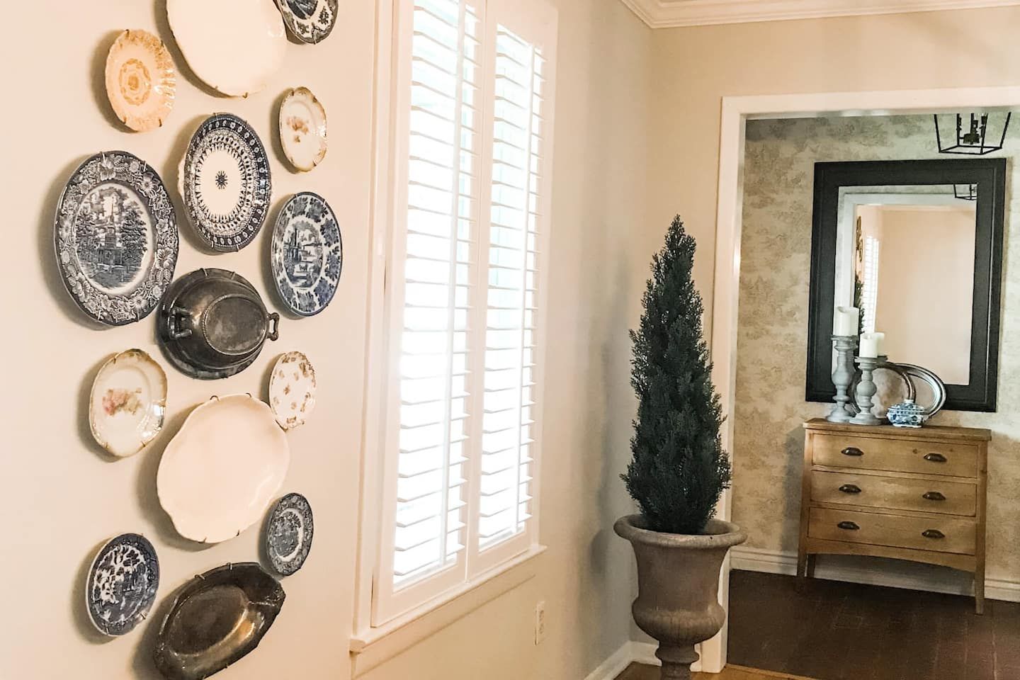 A room with a wall-mounted collection of plates, a potted evergreen, a mirror with a candle holder, and a wooden dresser.