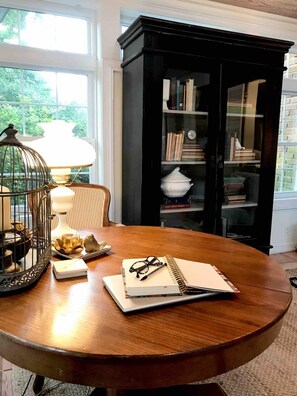 Een ronde houten tafel met een notitieboekje, brillen en een lamp. Achtergrond is een zwarte boekenkast met boeken en een witte pot.