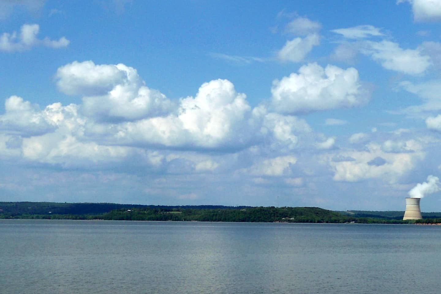 A body of water with a distant cooling tower emitting steam, surrounded by a hilly landscape under a partly cloudy sky.