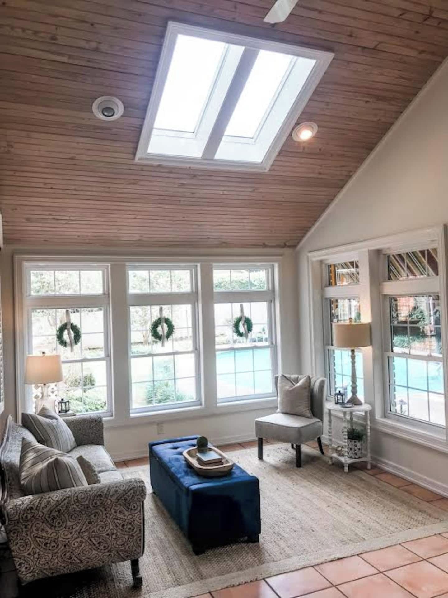 A room with a high ceiling, skylights, a patterned sofa, a blue ottoman, a chair, and a side table with a lamp.