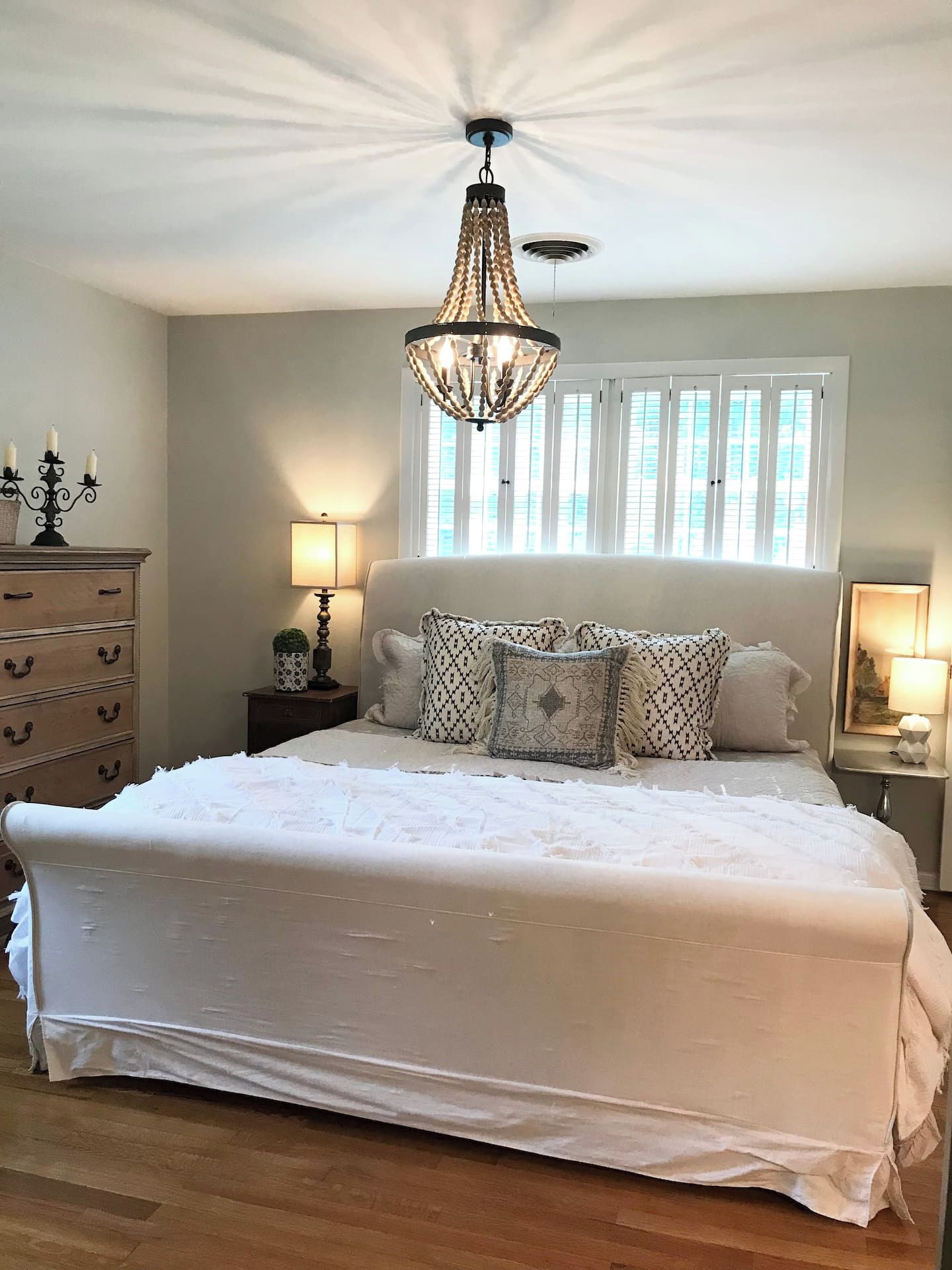 A bedroom with a large bed, a chandelier, a dresser, and a window with blinds.