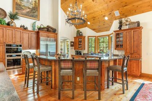 A spacious kitchen with wooden cabinets, a central island with bar stools, a chandelier, and a large window providing natural light. - Newly Listed! 8BR Beaver Creek Home | Hot Tub (Avon)