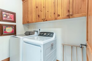 A laundry area with a front-loading washer and dryer, wooden cabinets above, and framed vintage advertisements on the wall. - Newly Listed! 8BR Beaver Creek Home | Hot Tub (Avon)