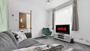 Een woonkamer met een grijze bank, een groene fauteuil, een televisie waarop 'NETFLIX' staat, en een deur die naar een andere kamer leidt.
