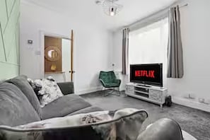 Uma sala de estar com um sofá cinza, uma poltrona verde, uma TV exibindo 'NETFLIX' e uma porta que dá para outro cômodo.