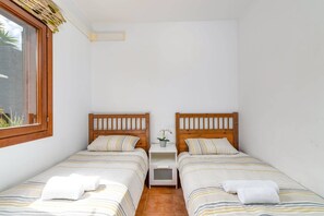 Ein kleines, sauberes Zimmer mit zwei Einzelbetten, einem Nachttisch mit Lampe und einem Fenster mit Blick auf Grünflächen.