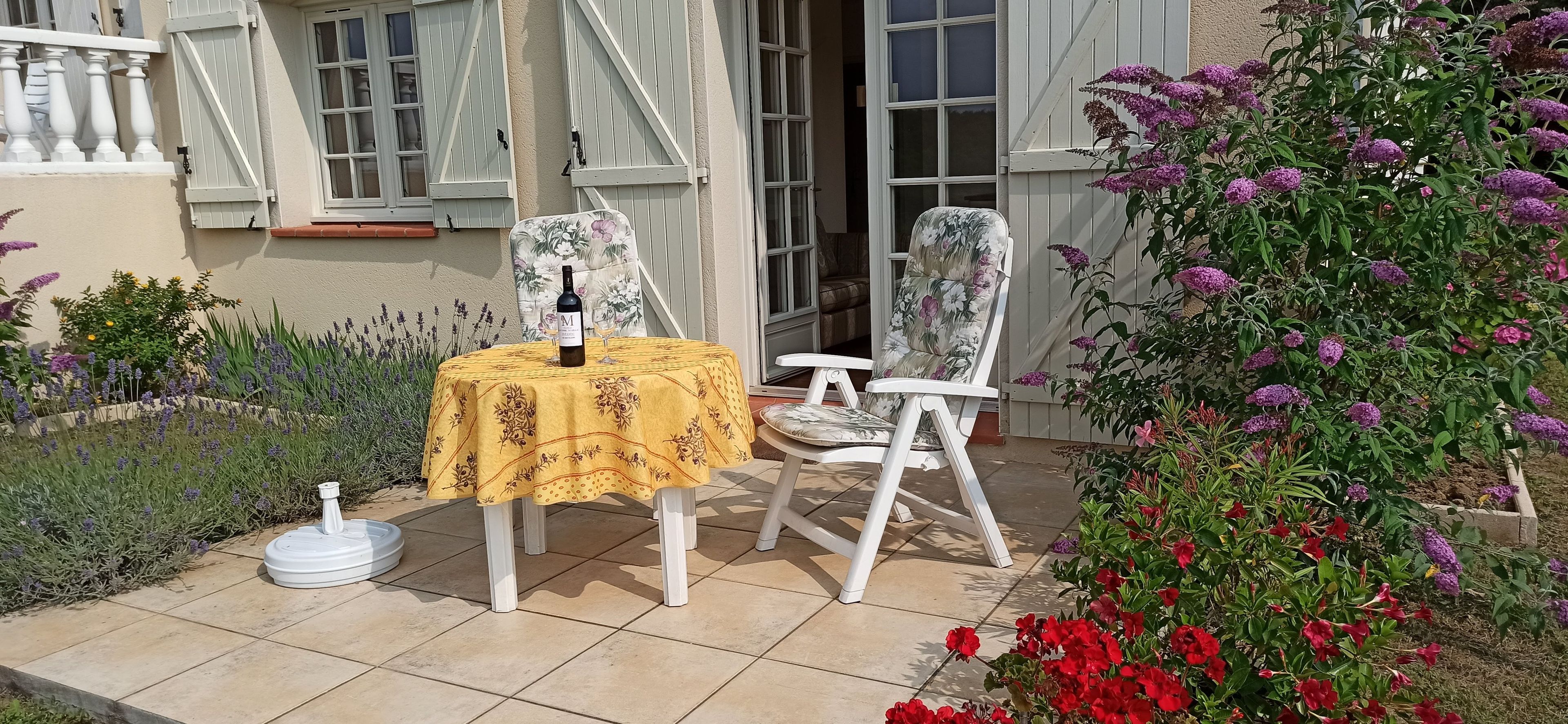 An outdoor patio with a table set for two, a bottle of wine, and two chairs with floral upholstery.