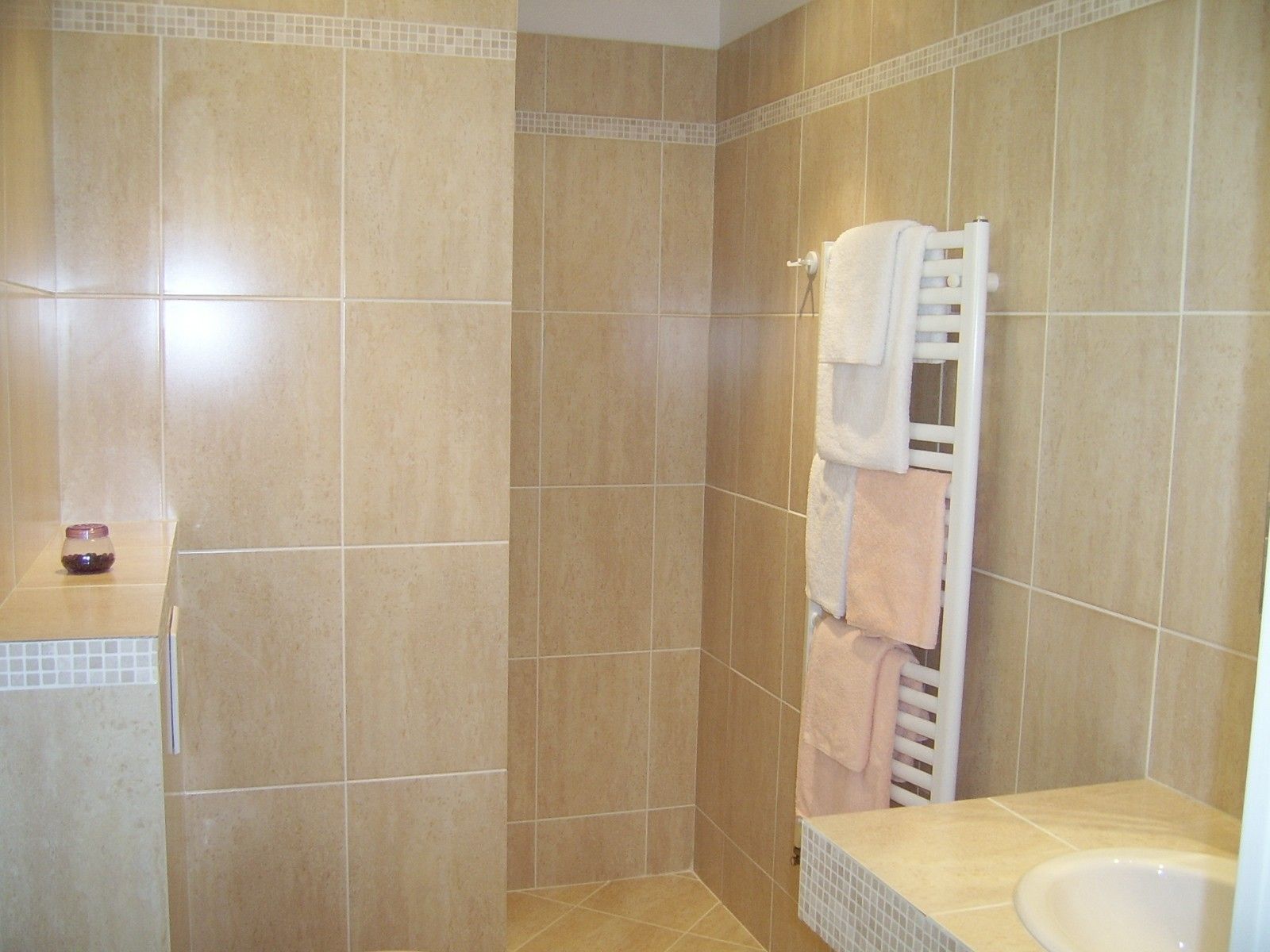 Un bagno con piastrelle color beige, un porta asciugamani bianco con asciugamani e un piccolo vasetto sul mobile.