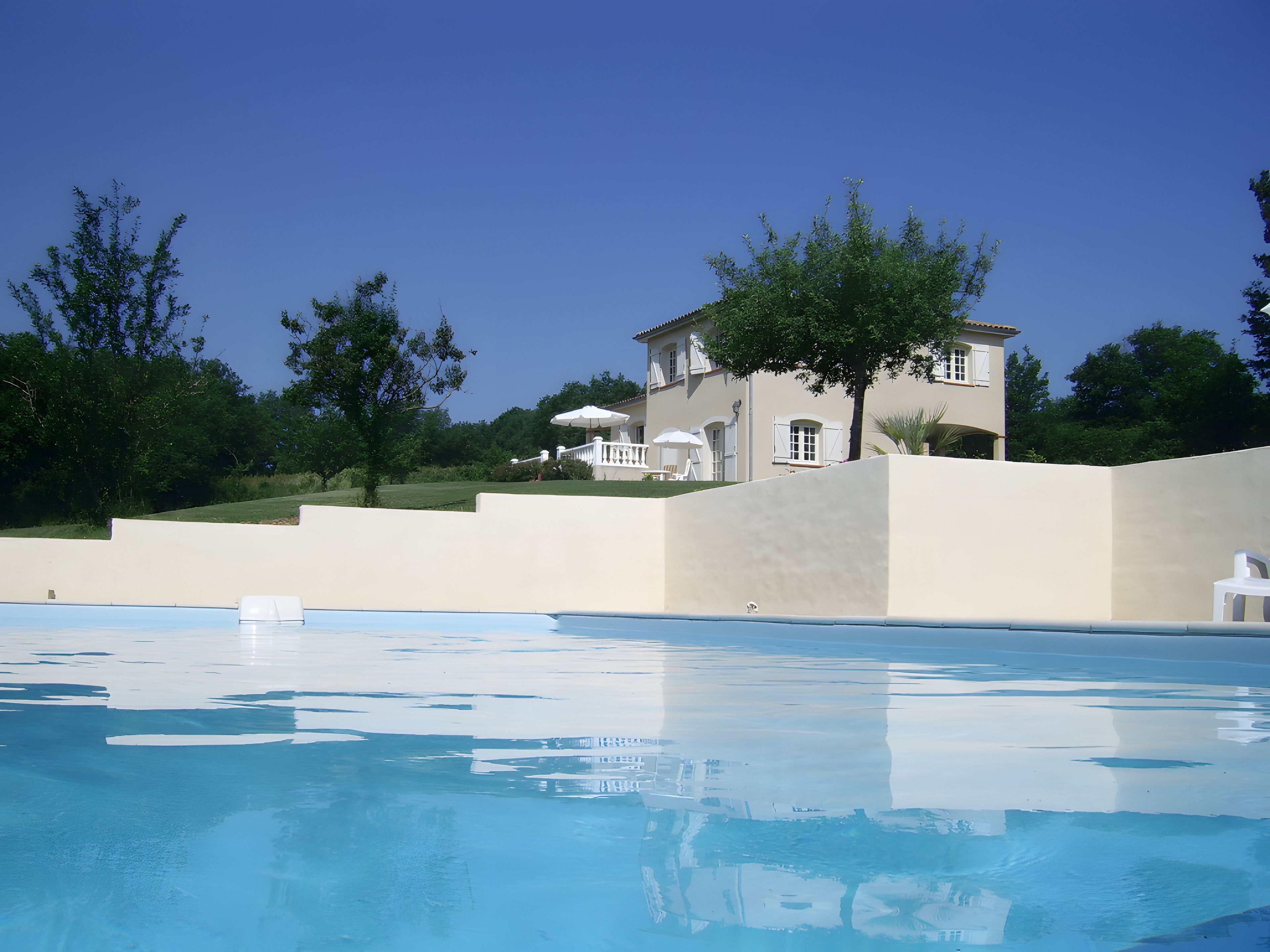 A swimming pool with a clear blue surface, a white building with multiple windows and balconies, and surrounding greenery.