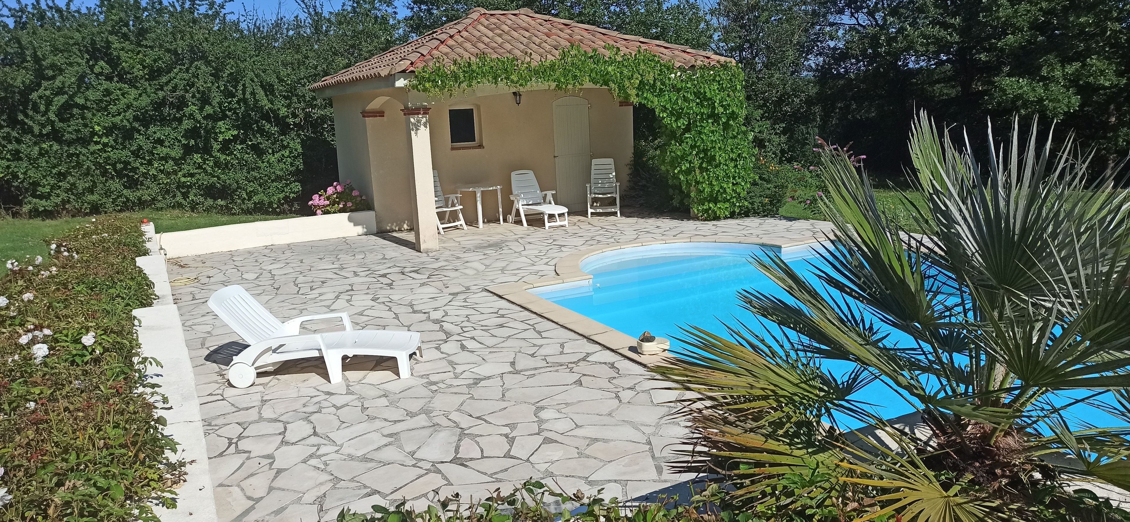 A pool area with stone flooring, lounge chairs, and a small building with a tiled roof.