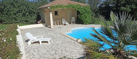 A pool area with stone flooring, lounge chairs, and a small building with a tiled roof.
