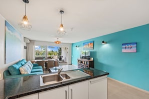 A modern kitchen with a large island, a dining area with a blue sofa, and a living room with a TV and artwork. - Tropical Paradise at Blue Bay | Pool & Beach Access (Willemstad)