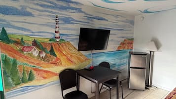 Habitación con un mural, televisión de pantalla plana, mesa con sillas, refrigerador y lámpara.