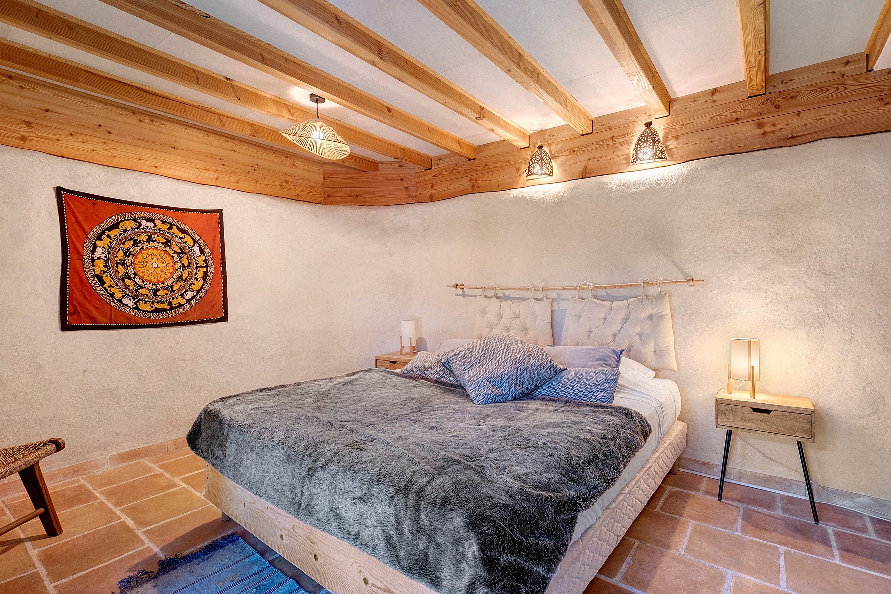 A bedroom with a wooden ceiling, a bed with a grey blanket, a nightstand with a lamp, and a tapestry on the wall.