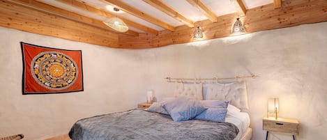 A bedroom with a wooden ceiling, a bed with a grey blanket, a nightstand with a lamp, and a tapestry on the wall.