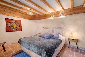 A bedroom with a wooden ceiling, a bed with a grey blanket, a nightstand with a lamp, and a tapestry on the wall. - Villa 'Sawubona' with Mountain View, Private Terrace and Wi-Fi (Les Adrets-de-l'Estérel)
