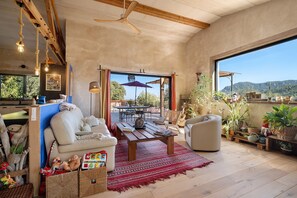 Un vaste espace de vie doté d’un plafond en bois, d’une grande fenêtre offrant une vue sur l’extérieur, d’un coin salon confortable avec un canapé et des fauteuils, et d’une table à manger avec des chaises.