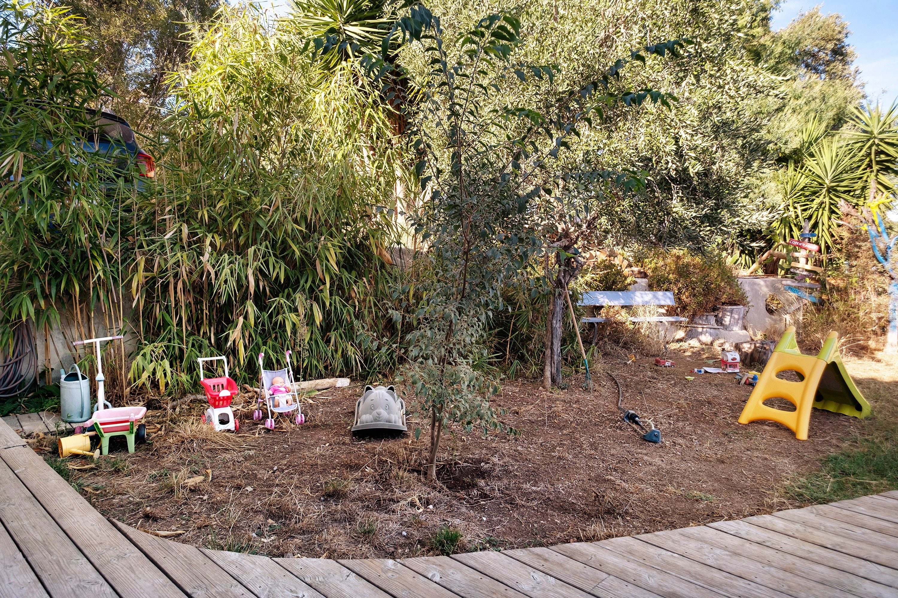 Une terrasse en bois avec un espace jardin comprenant un petit arbre, une voiture jouet et une poussette jouet.