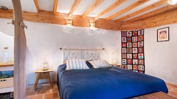 A bedroom with a bed, wooden beams, a wall tapestry, and a bedside table with a lamp.