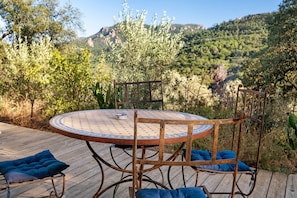 A round metal table with a patterned tabletop, surrounded by metal chairs with cushions, set on a wooden deck with a view of a mountainous landscape.